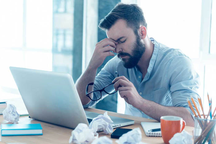 Frustrated young beard man massaging his nose and keeping eyes closed while sitting at his working place in office